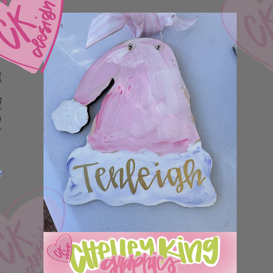 Adorable personalized hand painted wooden art ornament - Pink Santa Hat Approximately 6" ... Just let us know what you would like it to say, the color bow, and the color writing!:)

I design and personalize each of the ornaments to order!&nbsp; If you would like a different color hat or special design, please reach out to me at chelley@chelleyking.com!
